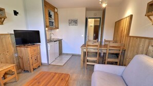 A cozy living room with a wooden coffee table, a sofa, and a TV. There's a kitchen with a microwave, sink, and cabinets. A dining table with chairs is also visible. - Comfortable 2-room, ski-in ski-out, near shops (La Tania)