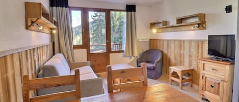A cozy room with a wooden dining table, two chairs, a sofa, a TV stand, and a window with curtains.
