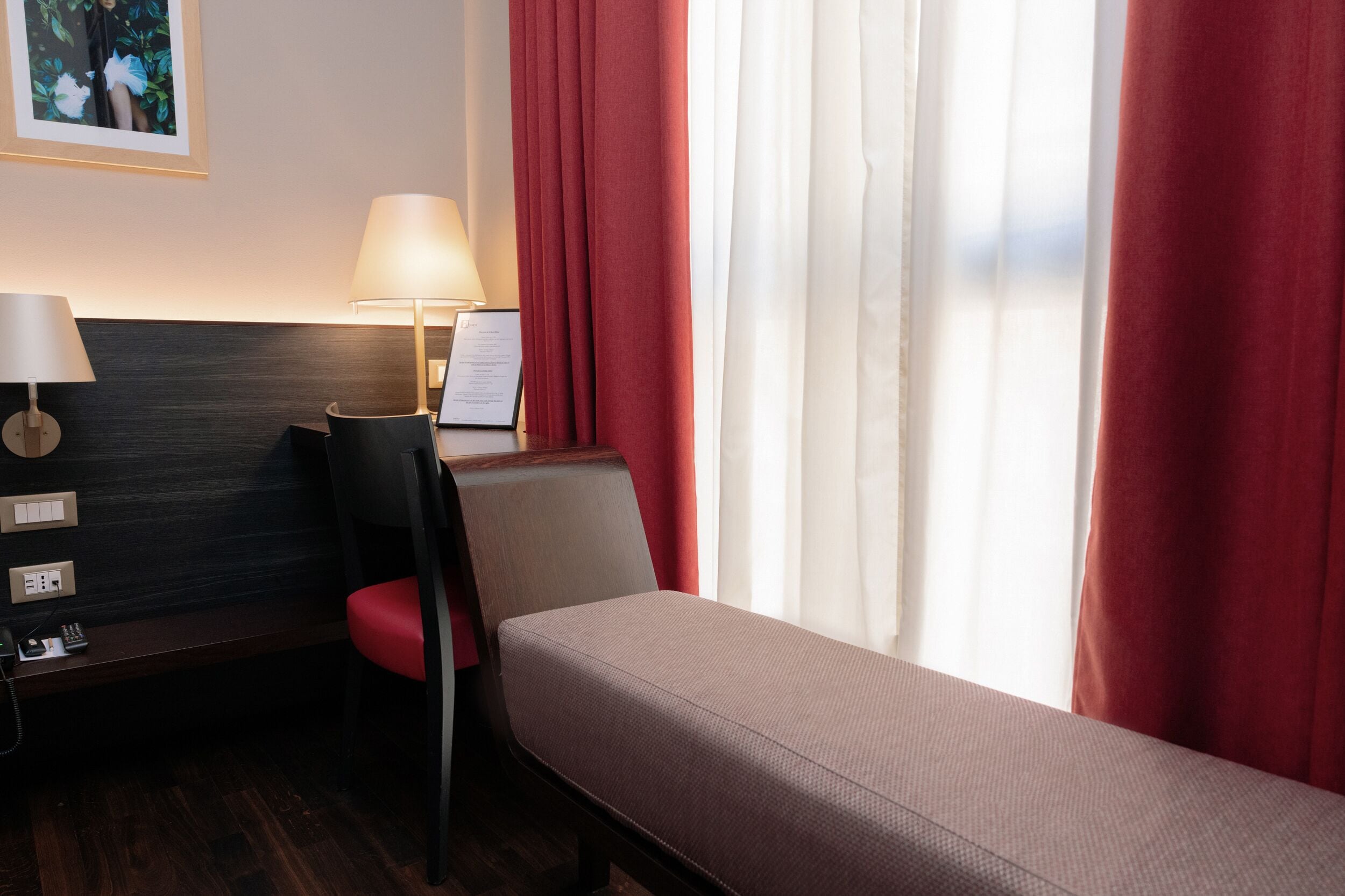 A hotel room with a single bed, a desk with a lamp, a chair, and a small table with a phone and a menu.