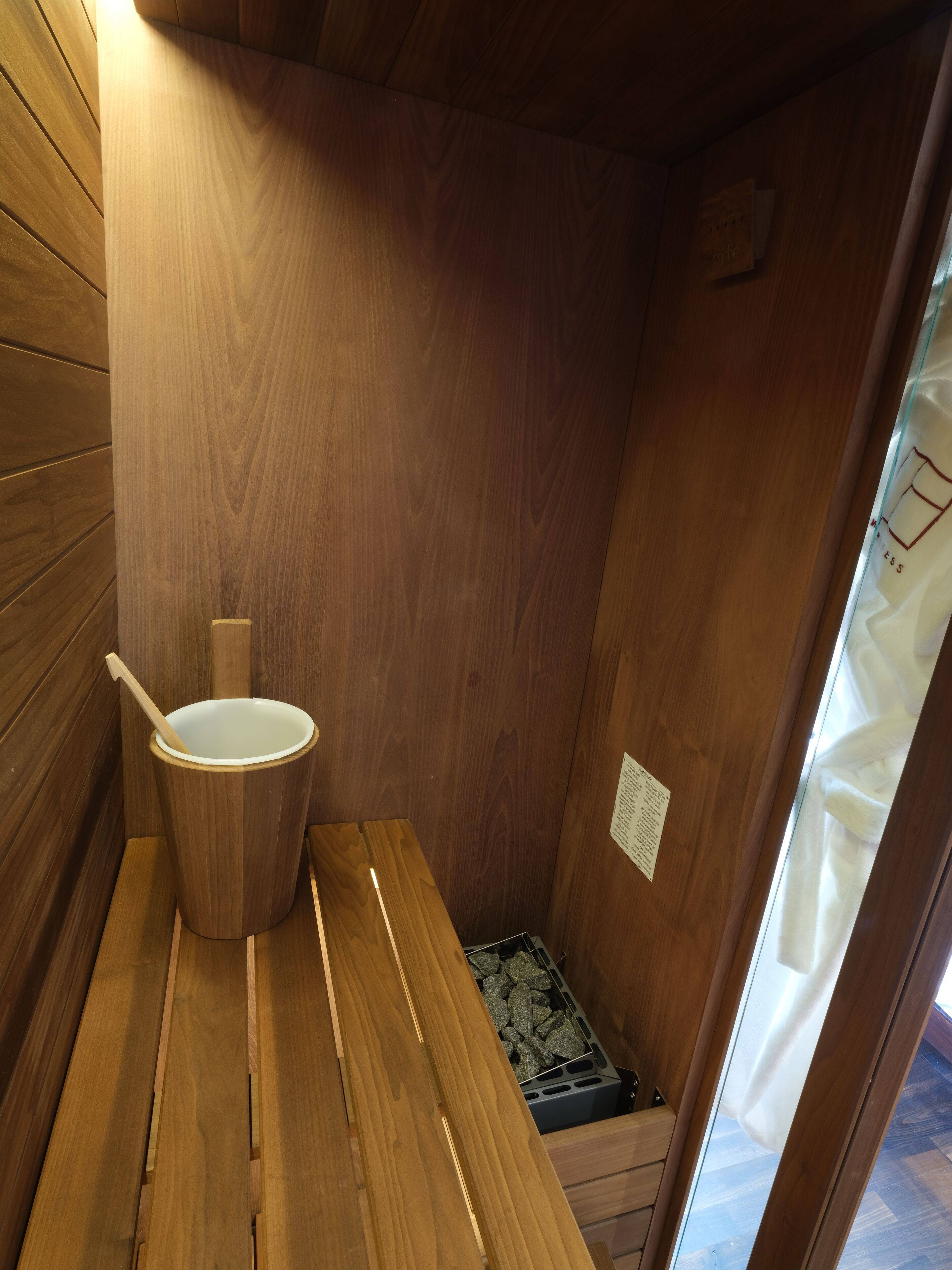 Un sauna en bois avec un banc en bois, un seau blanc doté d’une anse en bois et un petit plateau rempli de pierres.