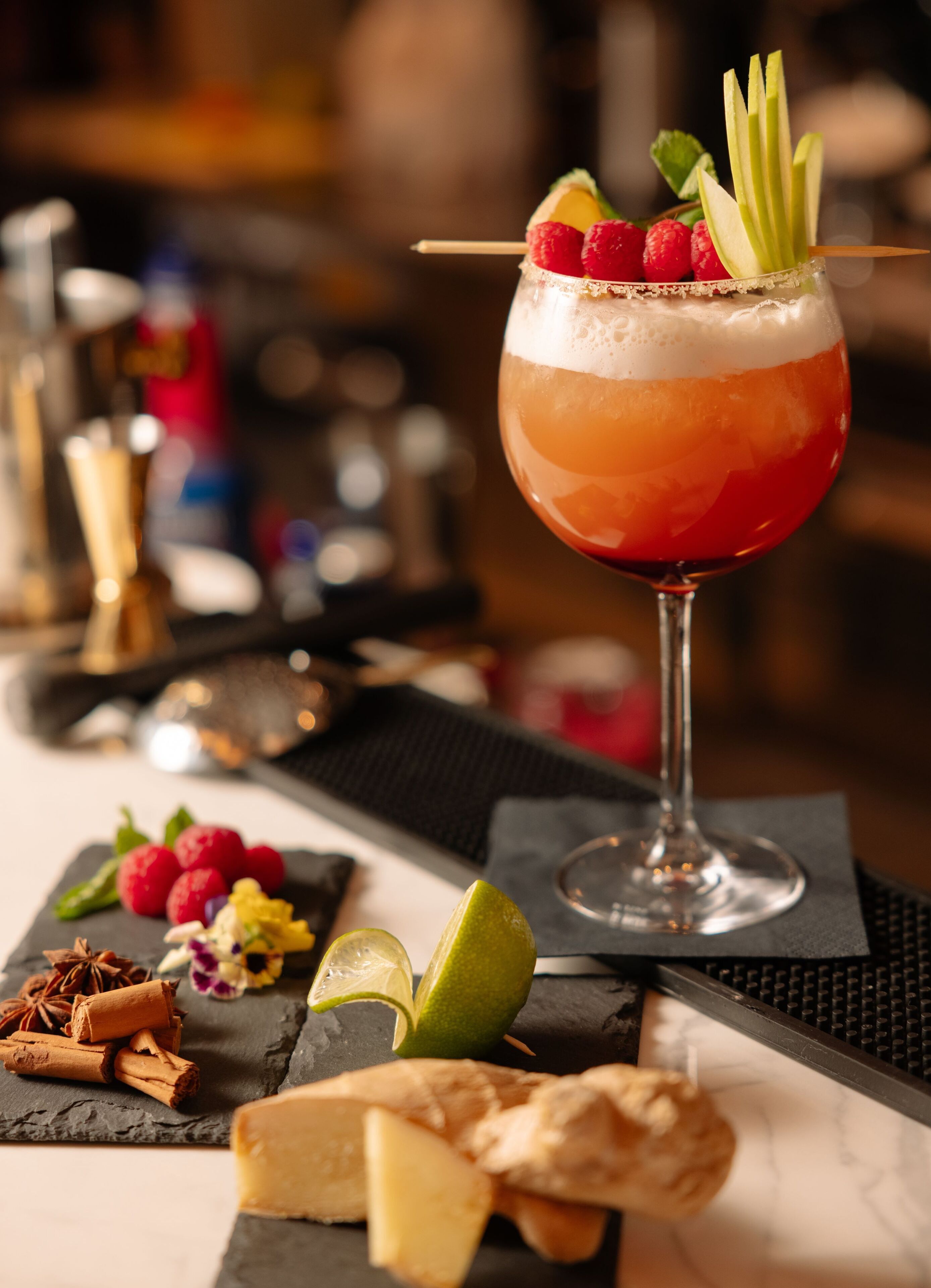 A cocktail with a layered drink, garnished with raspberries and apple slices, on a bar counter with a slate serving board, garnishes, and a glass of wine.