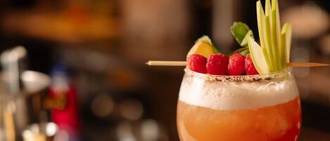 A cocktail with a layered drink, garnished with raspberries and apple slices, on a bar counter with a slate serving board, garnishes, and a glass of wine.