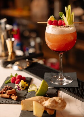 A cocktail with a layered drink, garnished with raspberries and apple slices, on a bar counter with a slate serving board, garnishes, and a glass of wine. - 10 KEYS MILANO    (Milan)
