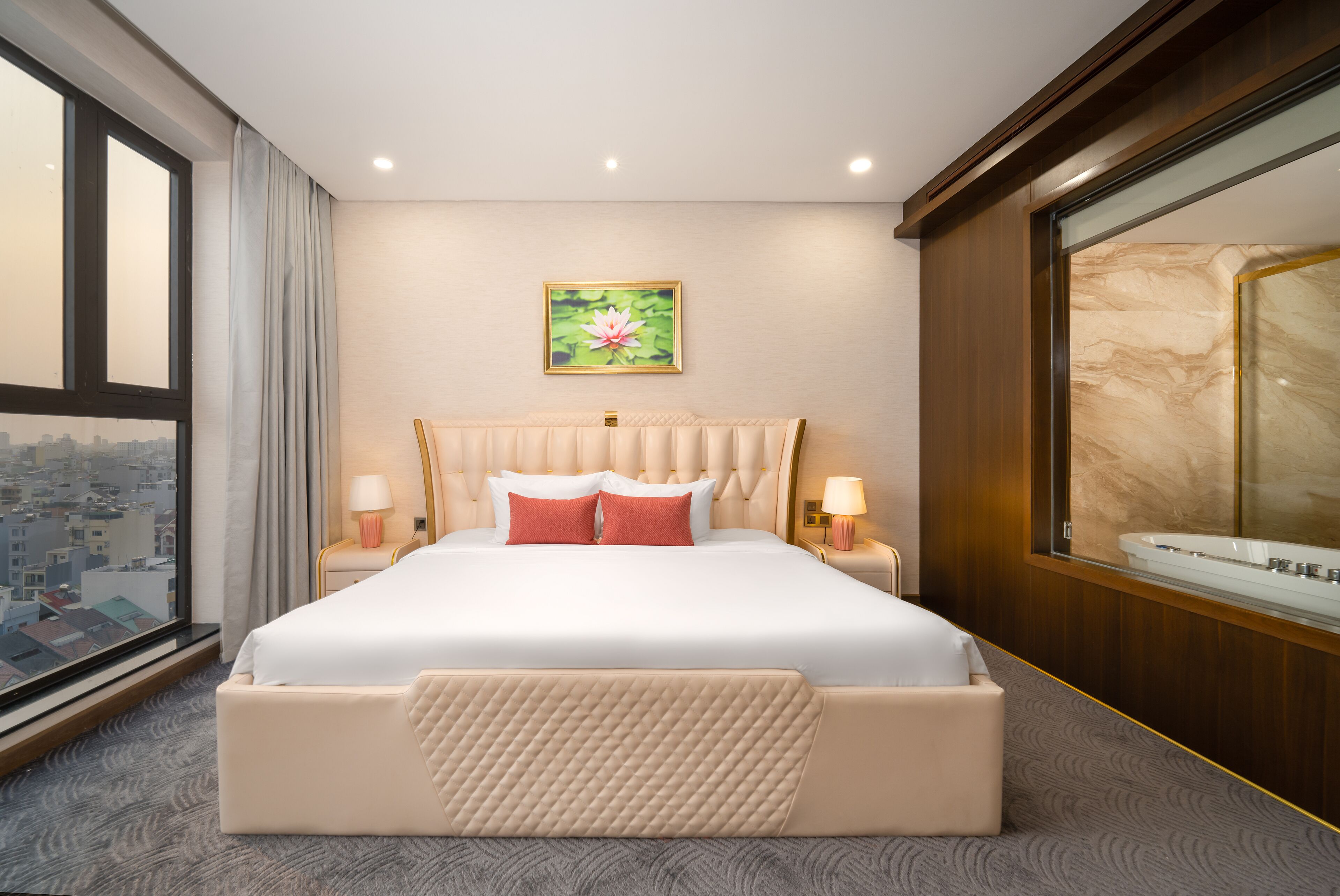 A hotel room with a large bed, a city view, a marble bathtub, and a painting of a lotus flower on the wall.