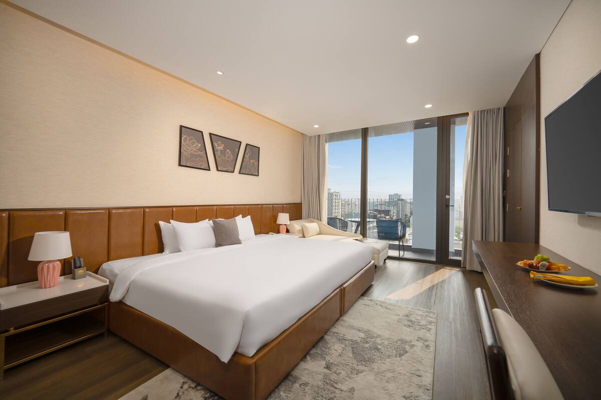 A hotel room with a large bed, a desk with a flat-screen TV, a nightstand, and a view of the city through large windows.