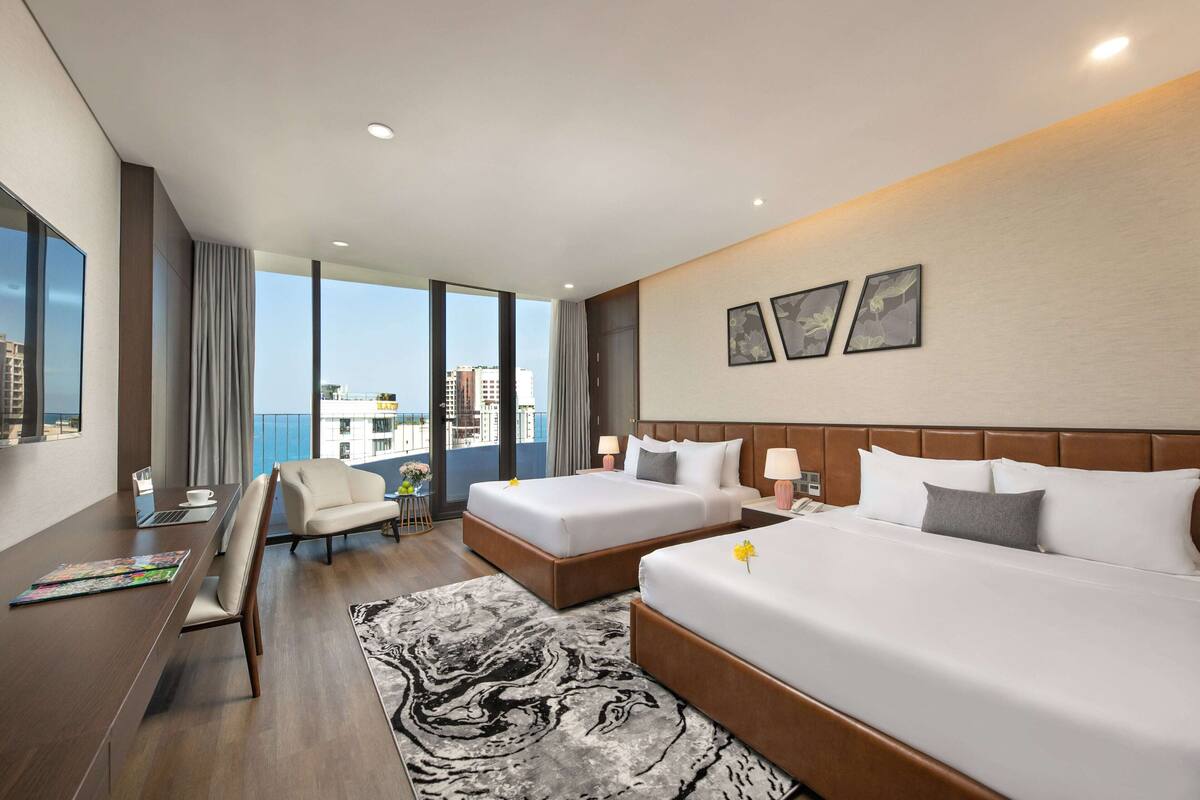 A hotel room with two beds, a desk, a chair, a large window, and a view of the city.