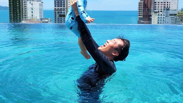 A man holding a child above water in a pool with a cityscape in the background. - Golden Lotus Grand (Da Nang)