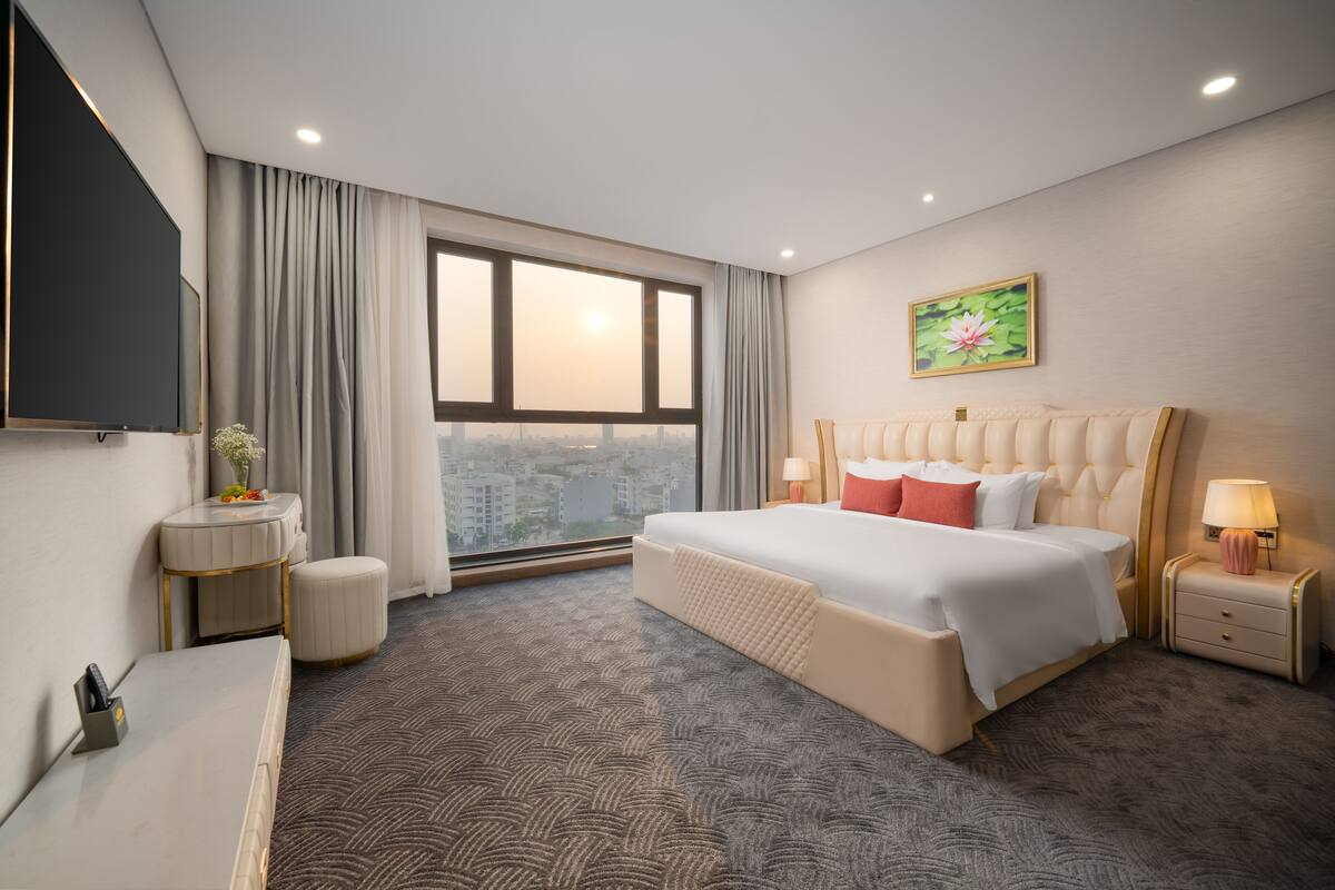A hotel room with a large bed, a desk, a chair, a small table, and a window with a city view.
