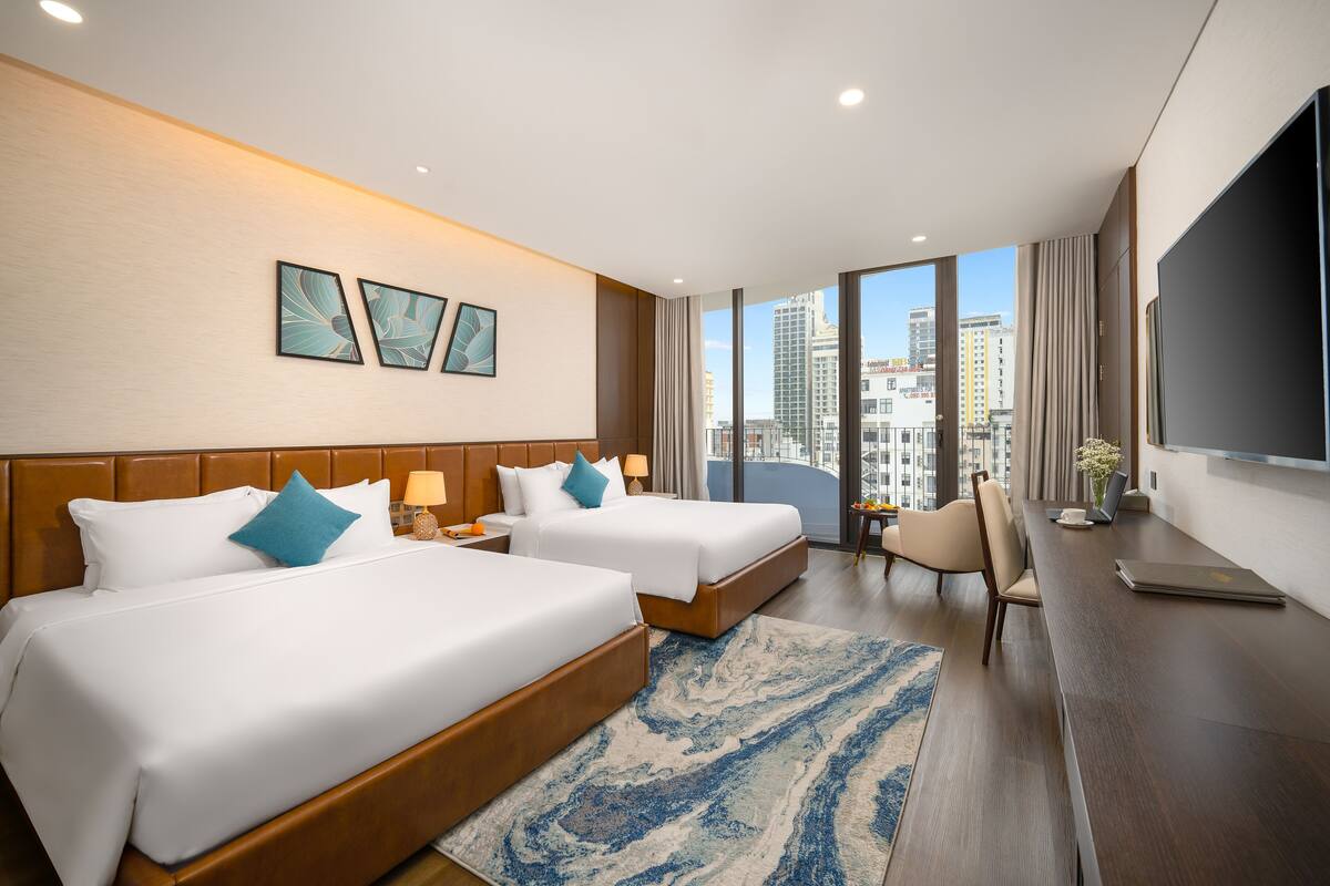 A hotel room with two beds, a desk, a chair, and a large window offering a city view.