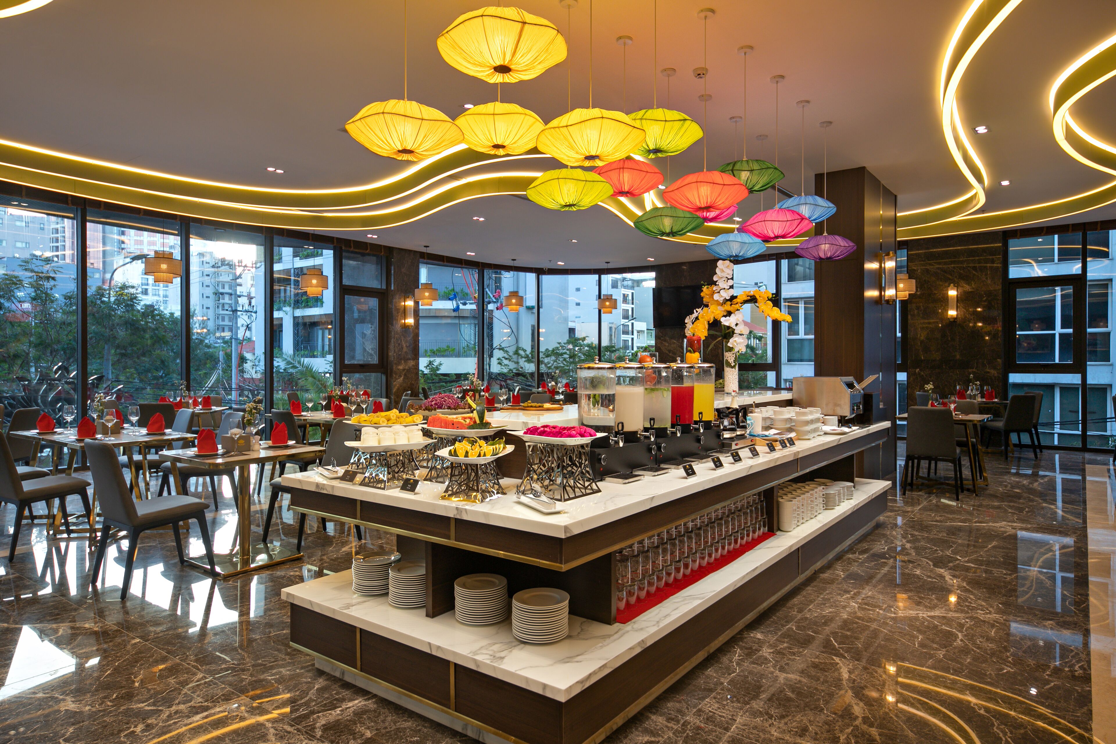 Un restaurante moderno con un bufé central, ventanas amplias y lámparas decorativas colgantes.