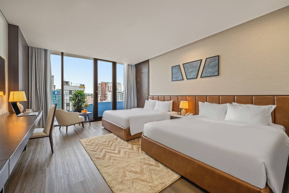 A hotel room with two beds, a desk, and a chair. There are large windows with a city view.