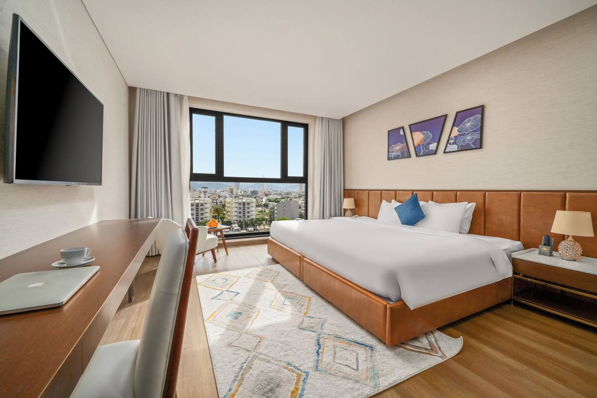 A hotel room with a large bed, a desk with a laptop, a TV, and a city view.