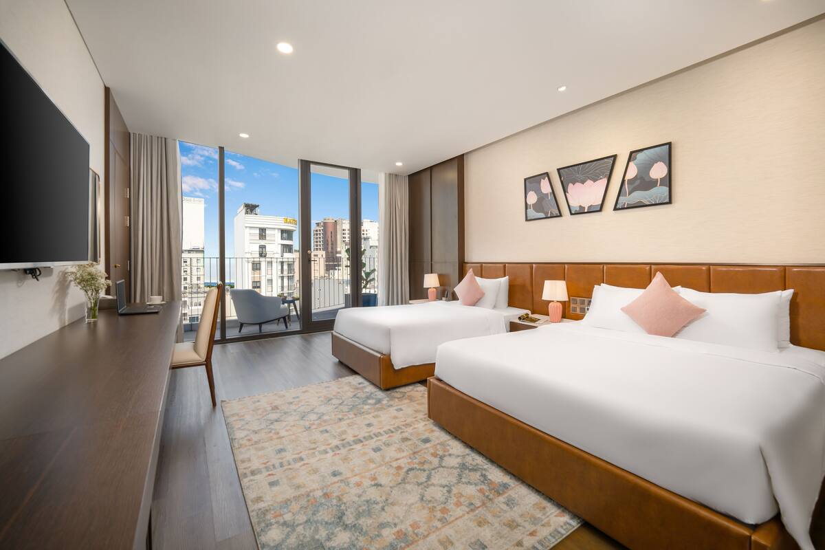 A hotel room with two beds, a desk, a TV, and a balcony with a city view.
