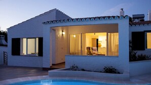 A modern house with a swimming pool, featuring a patio area with a chair and a table, and a visible interior with a lamp and curtains.