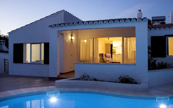 Uma casa moderna com piscina, apresentando uma área de varanda com cadeira e mesa, e interior visível com luminária e cortinas.