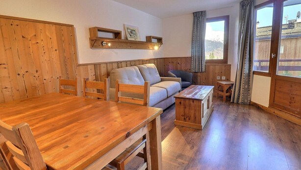 A cozy room with a wooden dining table, a sofa, and a small coffee table. There are wooden shelves above the sofa and a window with curtains. - Comfortable 2-Room + Alcove Apartment, Ski-in/Ski-out (La Tania)