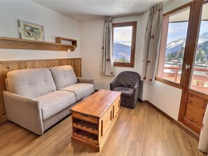 A room with a sofa, a chair, a wooden coffee table, a window with a mountain view, and a wooden shelf with a picture. - Comfortable 2-room apartment, ski-in/ski-out, near shops (La Tania)