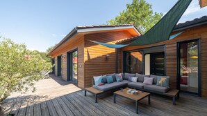 Eine Holzterrasse mit einer Couch und einem Tisch, großen Fenstern und einem grünen Blätterdach.