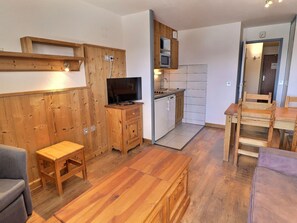 Ein kompakter Wohnraum mit einer Wandeinrichtung aus Holz, einem Fernseher, einer Küchenzeile mit Mikrowelle und einem Esstisch mit Stühlen.