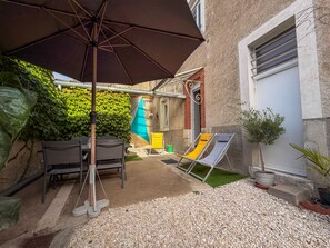 Een terras buiten met een tafel, stoelen, een gele loungestoel, een blauwe parasol en een pot met een plant.