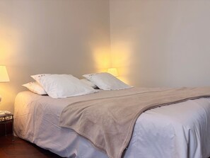 Un lit bien fait, avec une couverture beige et deux oreillers, une table de chevet avec une lampe et un luminaire fixé au mur.