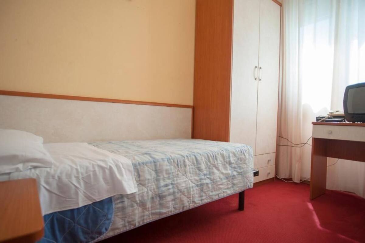 A small, single-bed room with a wooden headboard, a desk with a computer, and a wardrobe.