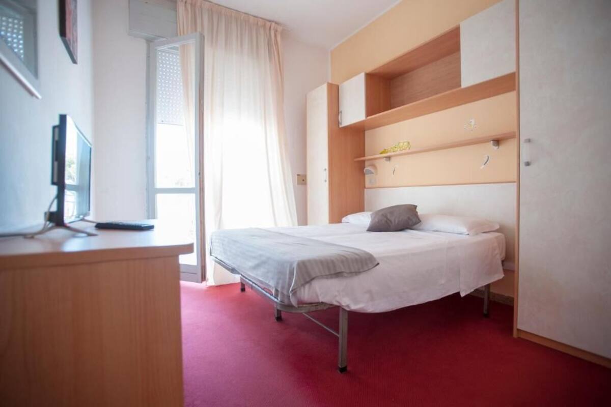 A bedroom with a bed, a desk with a computer, and a window with curtains.