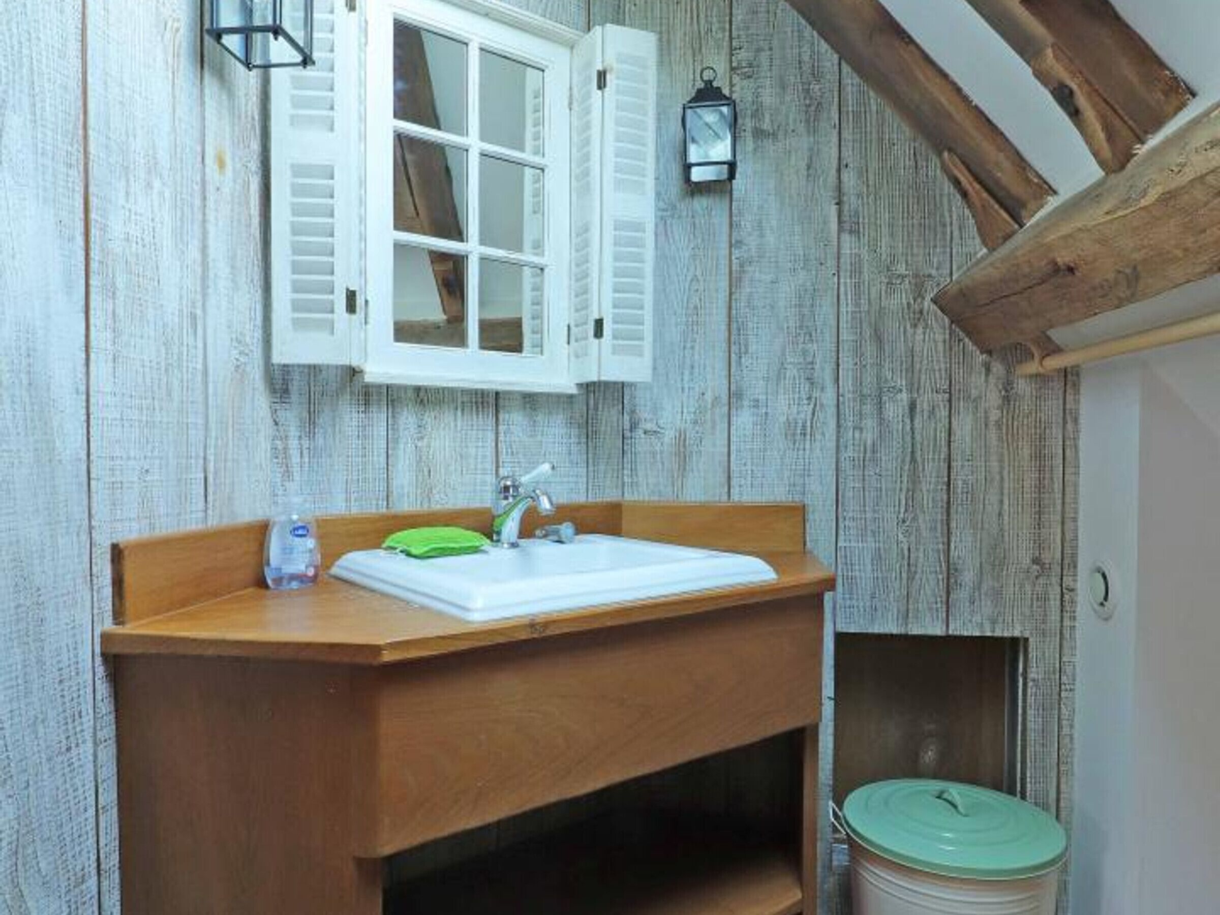 A bathroom with a wooden vanity, a white sink, and a green soap dispenser. There is a window with white shutters and a lantern on the wall.