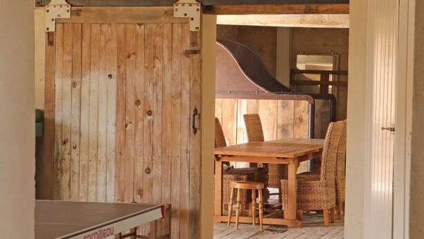 A rustic wooden room with a large sliding door, a wooden table, wicker chairs, and a grill in the background.