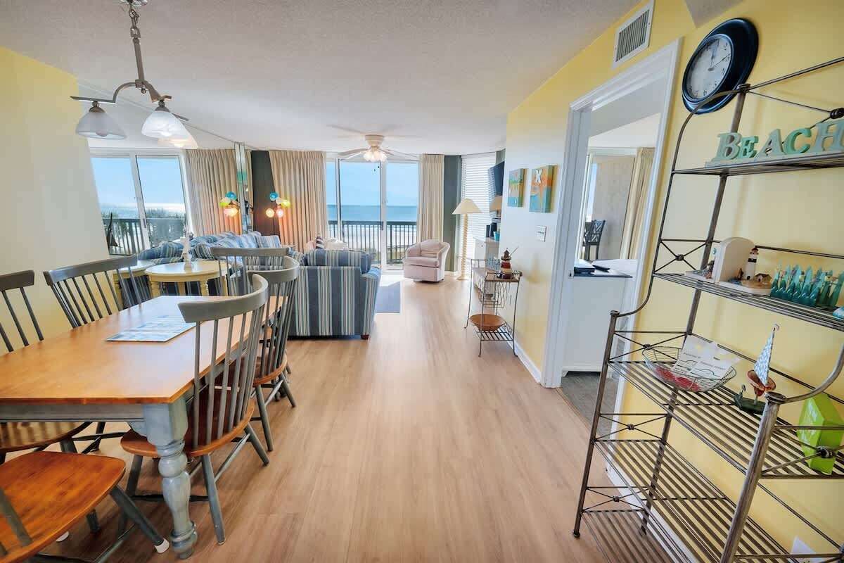 A hotel room with a dining area, a balcony with a view, and a shelf with items.