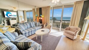 A spacious living room with a striped sofa, a round coffee table, and a beige armchair. A flat-screen TV is mounted on the wall, and a large window offers a view of the ocean. - New Private Ashworth Resort Direct Oceanfront View (North Myrtle Beach)