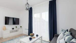 Una sala moderna con televisor de pantalla plana, mobiliario blanco, una mesa de centro con frutas y tazas, y un sofá con cojines estampados.