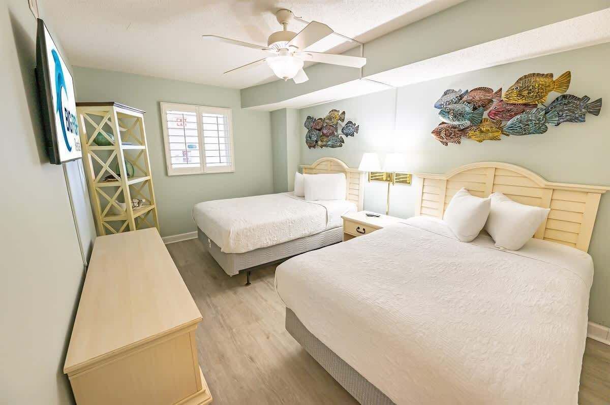 A bedroom with two beds, a ceiling fan, a TV, and artwork on the wall.