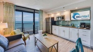 Une cuisine moderne avec des armoires blanches, un réfrigérateur en acier inoxydable et un four à micro-ondes. On y trouve un coin repas avec une table et des chaises, ainsi qu’un balcon offrant une vue sur l’océan.