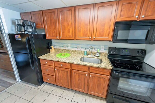 A kitchen with wooden cabinets, a granite countertop, a stainless steel refrigerator, a black microwave, and a black stove.