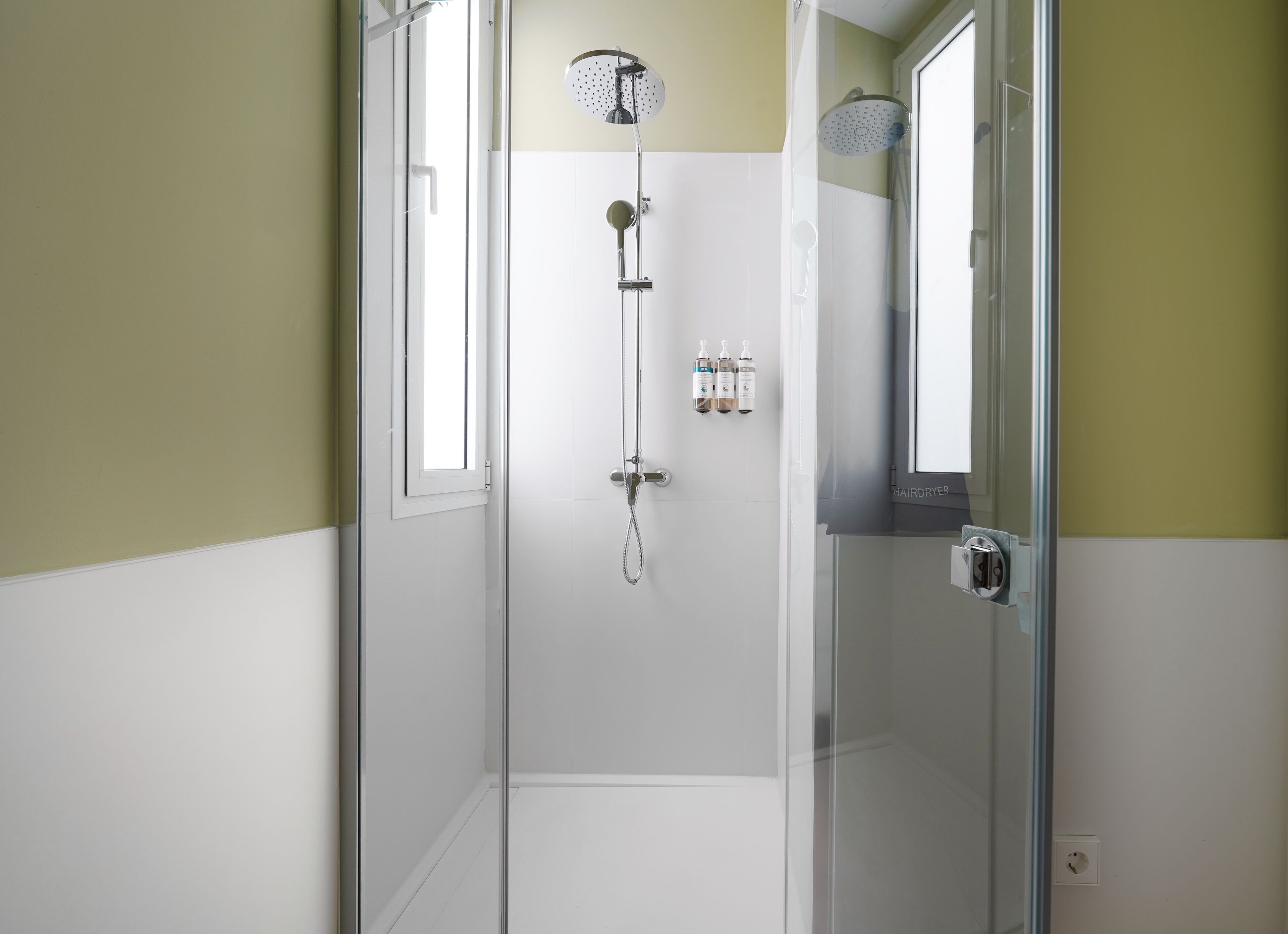 A glass-enclosed shower with a handheld showerhead and a wall-mounted showerhead. There are three bottles of shampoo and conditioner on the wall.
