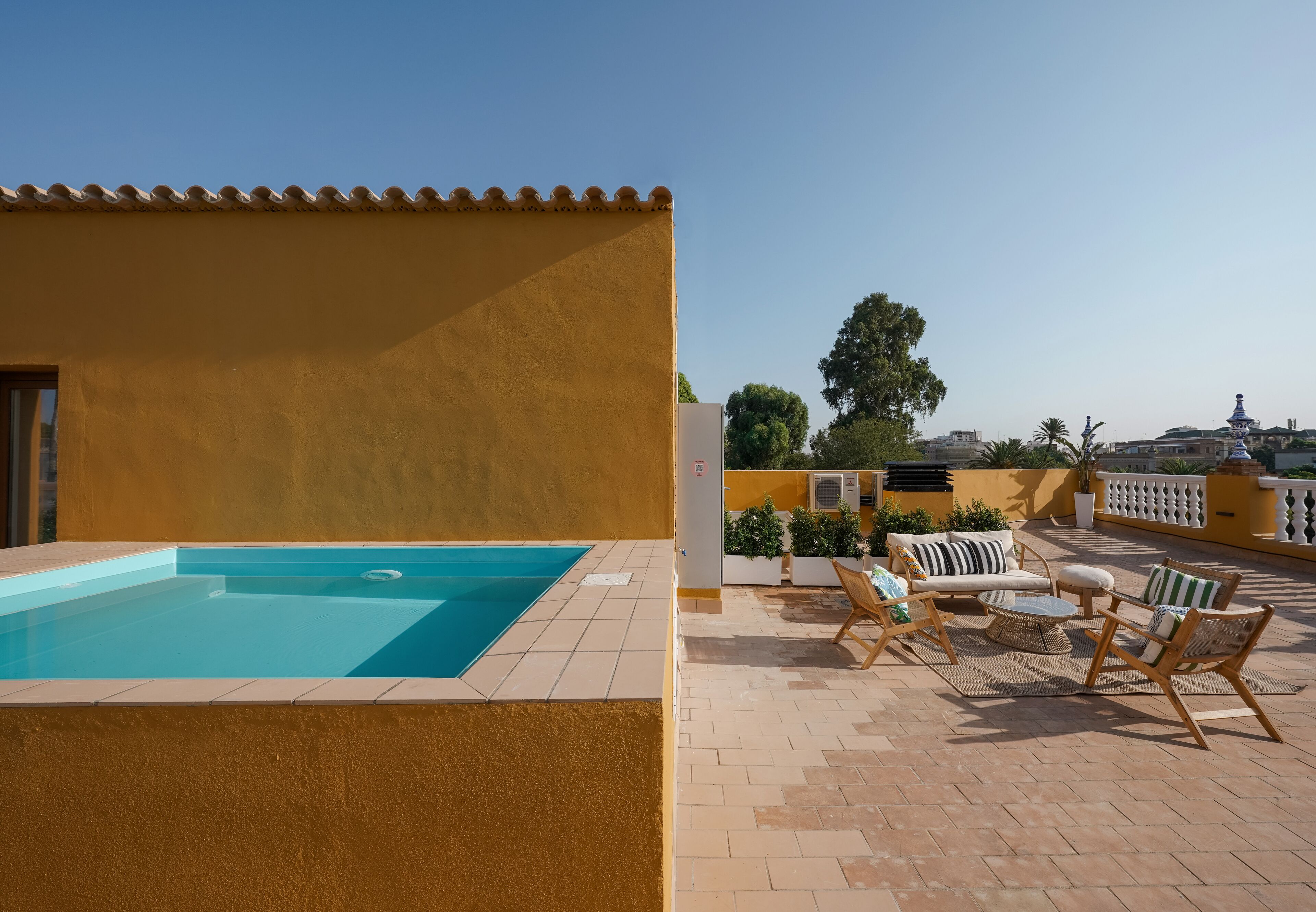 A swimming pool with a tiled deck, outdoor seating area with chairs and a table, and a view of the cityscape in the background.