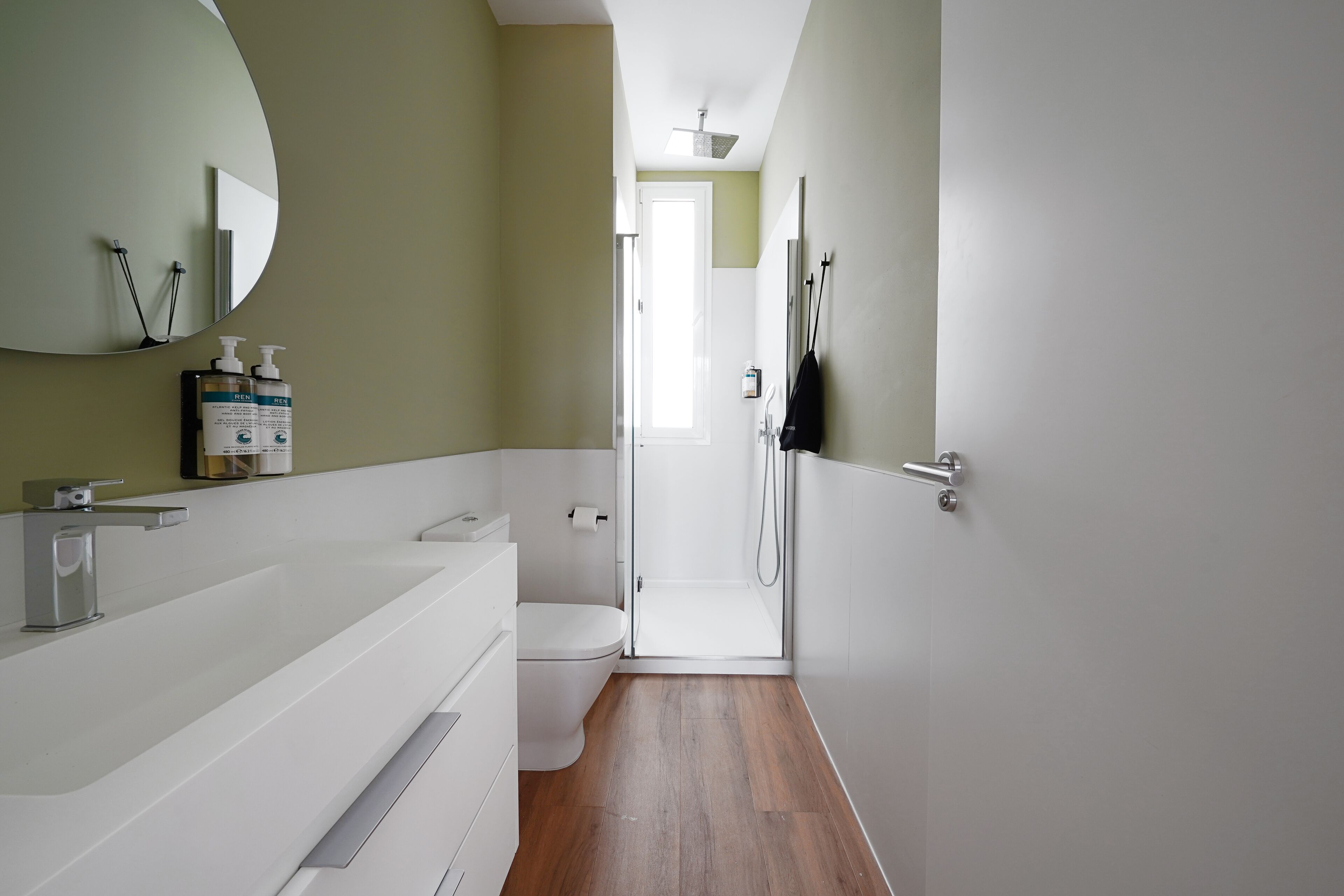 Un baño moderno con lavamanos, inodoro y ducha de color blanco. Hay artículos de aseo en el lavamanos y un espejo encima.