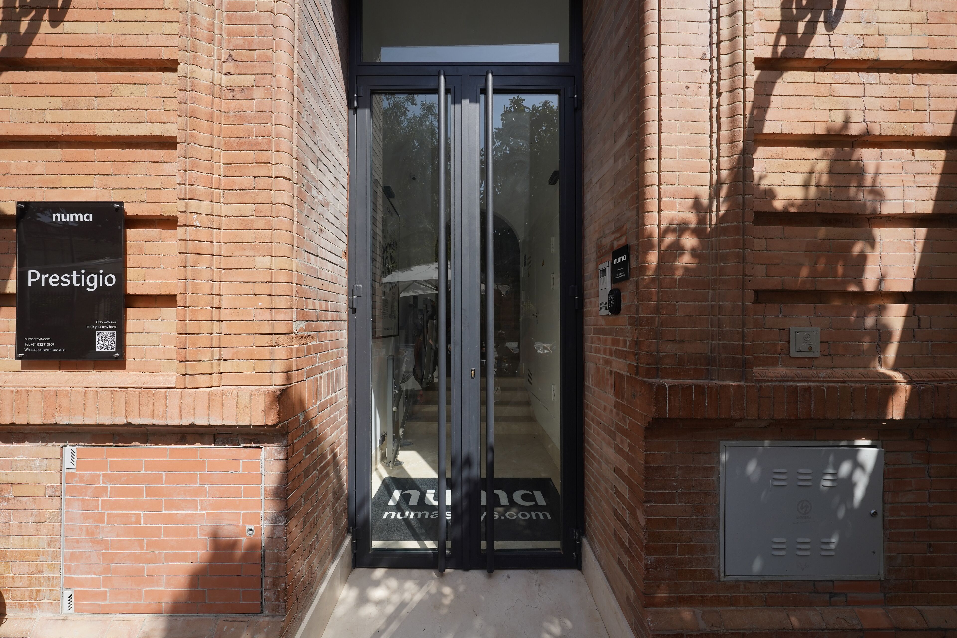 A brick building entrance with a glass door, a sign reading "numa Prestigio," and a doorbell.