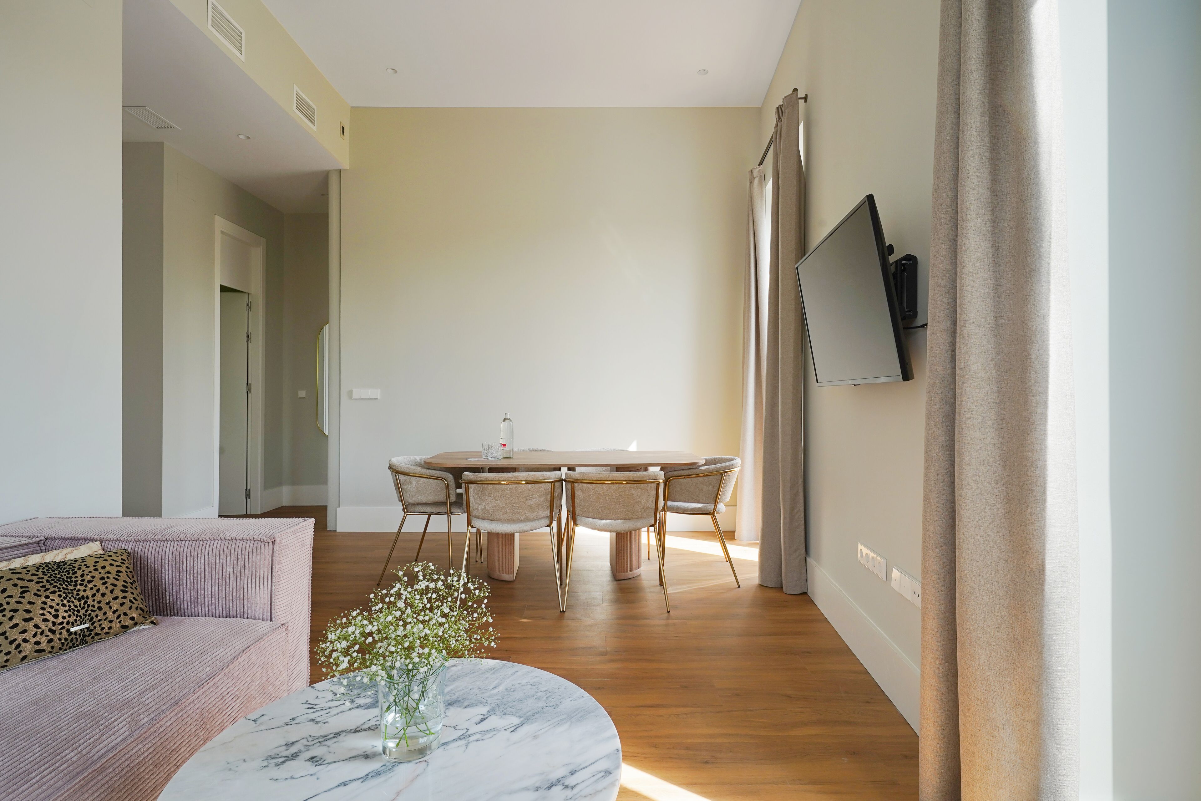 Ein modernes Wohnzimmer mit Sofa, rundem Couchtisch mit Vase, Essbereich mit Tisch und Stühlen sowie einem an der Wand montierten Fernseher.