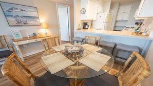 Una mesa redonda de vidrio con individuales tejidos, rodeada de sillas de mimbre con cojines oscuros, en una habitación con piso de madera y una zona de cocina al fondo.