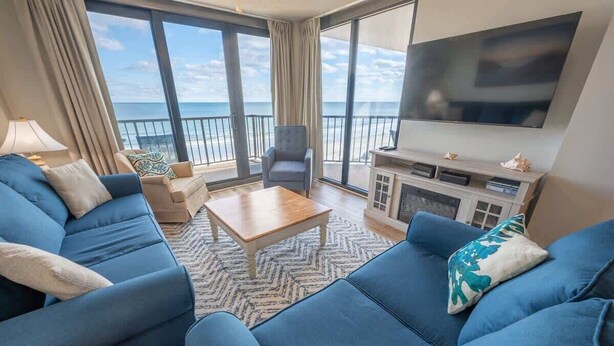 Un salón moderno con un televisor grande, asientos cómodos y un balcón con vistas al mar.