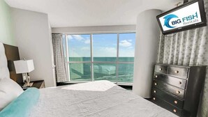 Camera d'albergo con un letto grande, una cassettiera, uno schermo televisivo che mostra 'Big Fish Rentals' e vista sull'oceano da una grande finestra.