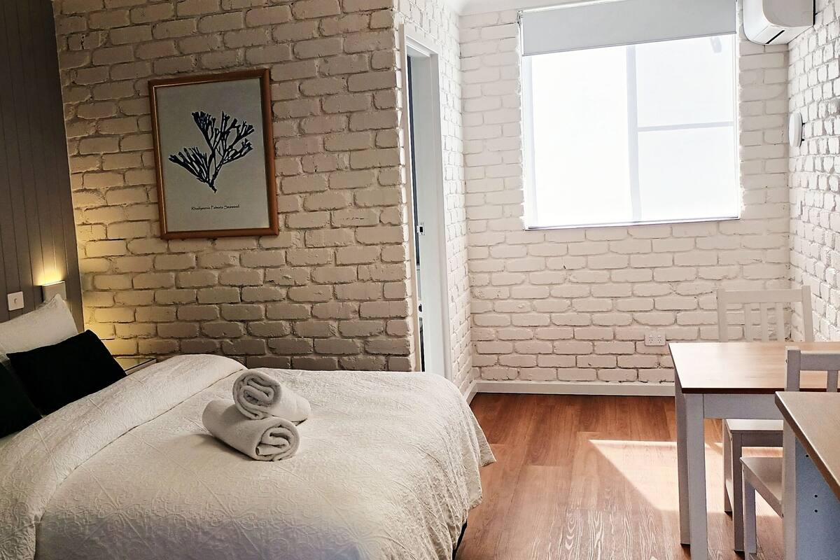 A bedroom with a bed, a framed botanical print, a window, and a wooden table.