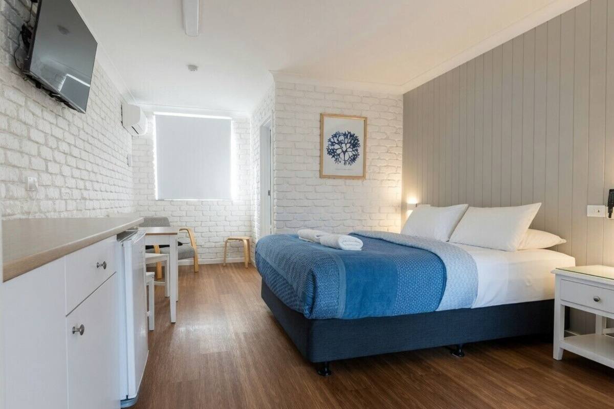 A bedroom with a bed, a desk, a chair, a TV mounted on the wall, and a window.