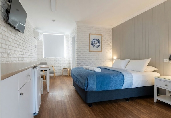 A bedroom with a bed, a desk, a chair, a TV mounted on the wall, and a window. - Bay House Motel (Bateau Bay)