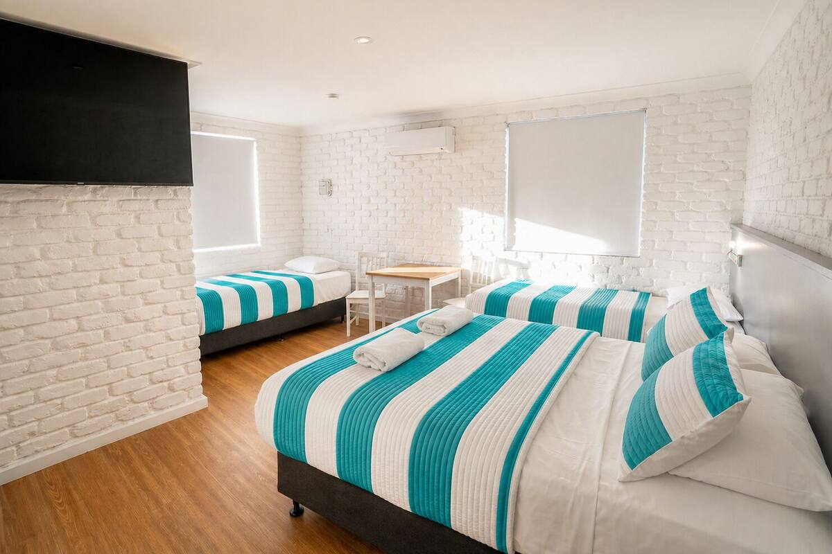 A hotel room with white brick walls, a wooden floor, two beds with striped bedding, a small table, and a TV mounted on the wall.