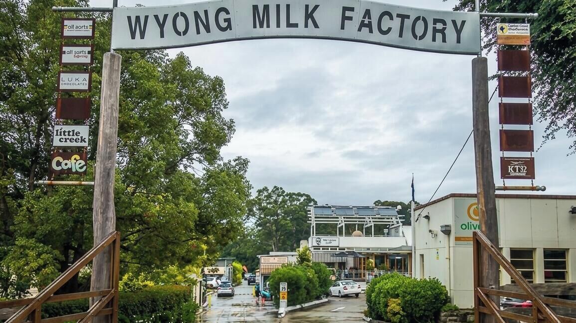 A sign for the Wyong Milk Factory with a wooden archway, a cafe sign, and a building in the background.