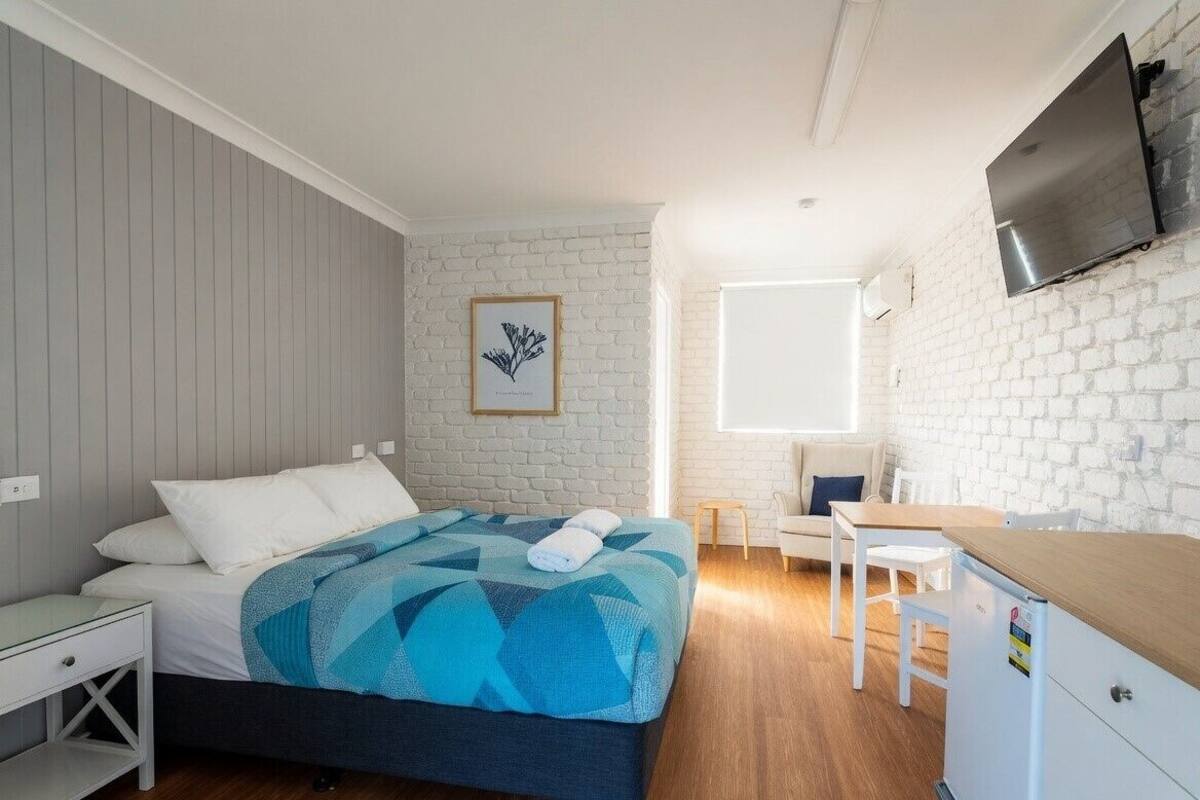 A bedroom with a bed, a desk, a chair, a TV, and a window.