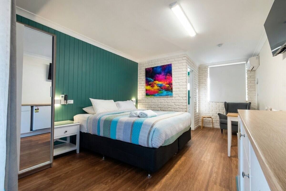 A bedroom with a bed, a desk, a chair, a wardrobe, and a colorful painting on the wall.